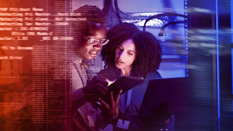 Two developers in a data center examine strings of code on a tablet and a laptop, working on a software development project.