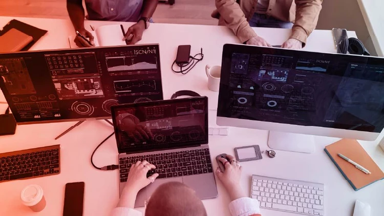 Team impegnato nel software development, con codici, dati e interfacce di analisi visualizzati su laptop e monitor multipli.